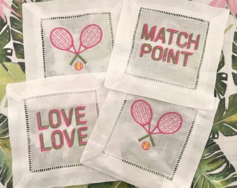May include: Four white linen napkins with pink and green embroidery. Two napkins feature tennis rackets and a tennis ball. One napkin says "LOVE LOVE" and the other says "MATCH POINT".