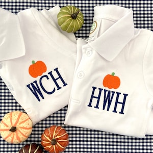 May include: Two white polo shirts with embroidered pumpkins and personalized monograms. The shirts are laid out on a blue and white gingham fabric with small decorative pumpkins.