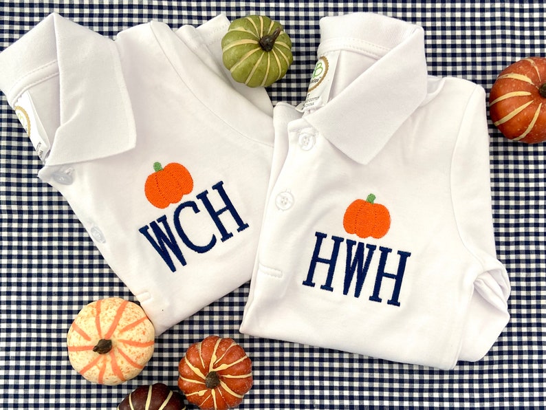 May include: Two white polo shirts with embroidered pumpkins and personalized monograms. The shirts are laid out on a blue and white gingham fabric with small decorative pumpkins.