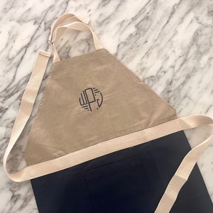 以下が含まれることがあります： 白いストラップが付いたベージュと黒の エプロン。エプロンにはポケットと、黒い文字で「WPJ」とモノグラムされた円形があります。