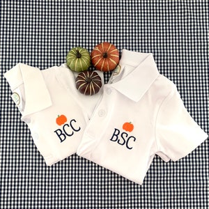 May include: Two white short-sleeved polo shirts with embroidered pumpkins and the letters "BCC" and "BSC" on a black and white gingham background.