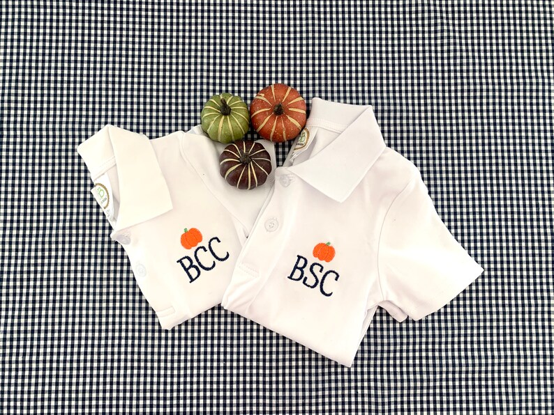 May include: Two white short-sleeved polo shirts with embroidered pumpkins and the letters "BCC" and "BSC" on a black and white gingham background.