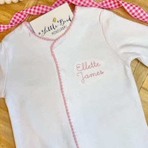 Puede incluir: Mono blanco para bebé con ribete rosa y el nombre bordado "Ellette James". El mono tiene mangas largas y cierre de broches. Una etiqueta con el texto "Little Bird Monograms" es visible.