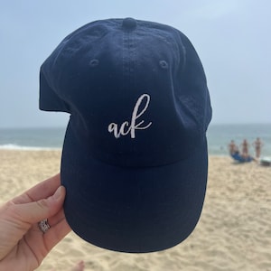 Può includere: Un cappello da baseball blu navy con la parola "ack" ricamata in bianco sul davanti.