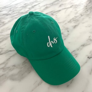 Pode incluir: Boné de beisebol verde com aba curva. O boné apresenta as letras "chs" bordadas em branco na frente. O chapéu é feito de um tecido macio e tem um botão na parte superior.