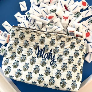 Puede incluir: Una bolsa acolchada blanca con un estampado floral azul y el nombre "Mahy" bordado en azul. La bolsa está llena de fichas de Mahjong blancas con marcas azules y rojas, esparcidas sobre una superficie azul.
