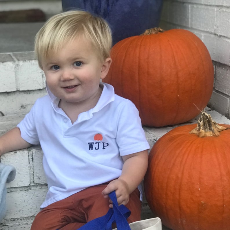 May include: A young child wearing a white polo shirt with an embroidered pumpkin and the letters "WJP" sits on a brick step with two large orange pumpkins.