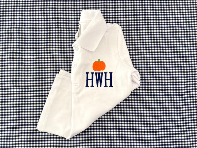 May include: White baby onesie with a pumpkin and the monogram HWH embroidered on the front. The onesie is lying on a black and white gingham fabric.