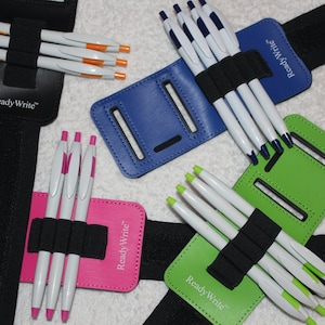 Może przedstawiać: Cztery kolorowe opaski na nadgarstek z czarnymi elastycznymi paskami i wieloma uchwytami na długopisy. Każda opaska ma nadrukowany tekst "Ready Write". Opaski są różowe, niebieskie, zielone i czarne.