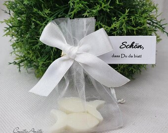 Könnte beinhalten: Ein weißer Organzabändchenbeutel mit einer weißen Satin Schleife gebunden. Der Beutel enthält zwei weiße, fischförmige Seifen. Eine weiße Karte mit schwarzem Text lautet "Schön, dass Du da bist!"