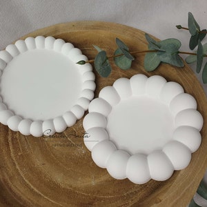 Può includere: Due sottobicchieri in ceramica bianca con un design a bordo rialzato e arrotondato. I sottobicchieri sono su una superficie in legno.