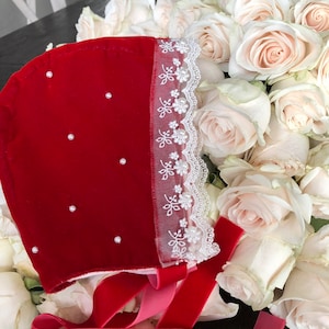 May include: A red velvet bonnet with white lace trim and pearl accents. The bonnet is tied with a red velvet ribbon and a pink ribbon.