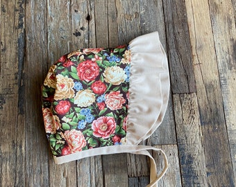May include: A floral bonnet with a brown and white floral print and a beige ruffle trim. The bonnet has two long beige ties.