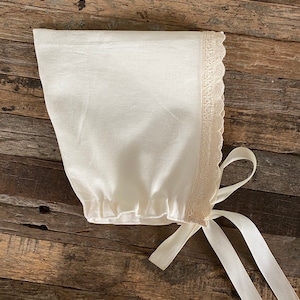 May include: Ivory-colored baby bonnet with a delicate lace trim along the edge. The bonnet is made of a soft fabric and features long, white ribbon ties. The bonnet is designed to be tied under the chin.
