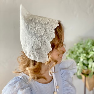 Puede incluir: Un gorro de encaje blanco con bordado floral. El gorro lo lleva una persona que viste un vestido a rayas azules y blancas. El gorro tiene un borde festoneado y está atado con una cinta blanca.