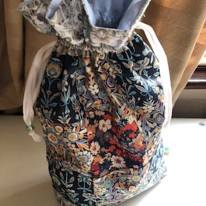 May include: A drawstring bag with a floral pattern in navy, orange, and cream. The bag's interior is light blue, and it has white drawstrings with decorative beads. The fabric has a detailed botanical design.