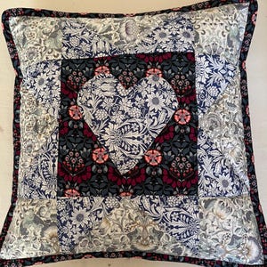 May include: Square cushion with a patchwork design. The cushion features a central heart shape and floral patterns in shades of blue, white, grey, and burgundy. The edges are trimmed with a dark blue border.