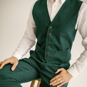 Pode incluir: Um conjunto verde escuro composto por um colete, calças a condizer e uma camisa branca abotoada. O colete tem decote em V, bolsos e botões castanhos. O conjunto é feito de tecido texturizado, possivelmente linho. O estilo é clássico e elegante.