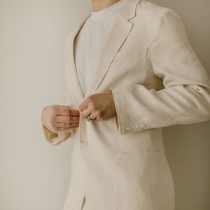 Puede incluir: Un traje beige claro, compuesto por una americana y un pantalón, se está abrochando. La americana tiene solapas con muescas, tres botones en la manga y un bolsillo en el pecho. Se lleva una camiseta blanca debajo. El traje es de tejido ligero y texturizado.