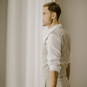 Pu&ograve; includere: Un uomo con una camicia bianca e un gilet beige si trova davanti a una tenda bianca. Sta guardando a destra del fotogramma.