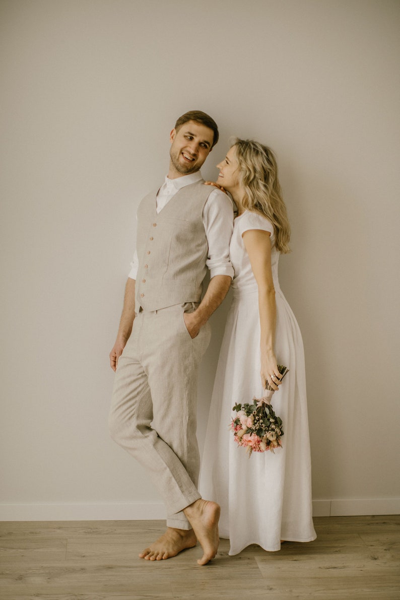 Pu&ograve; includere: Una coppia in una stanza bianca, la donna indossa un abito bianco e tiene un mazzo di fiori, l'uomo indossa un gilet beige e pantaloni, si guardano e sorridono.