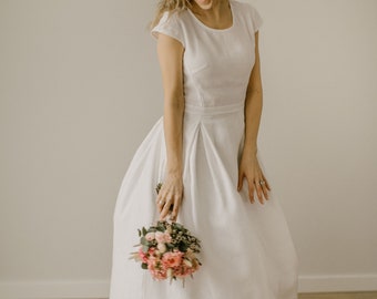 May include: A woman in a white linen dress smiles while holding a bouquet of pink and green flowers.