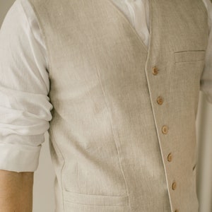Pu&ograve; includere: Un gilet di lino beige chiaro indossato su una camicia bianca con colletto. Il gilet ha una tasca sul lato sinistro e sei bottoni sul davanti.