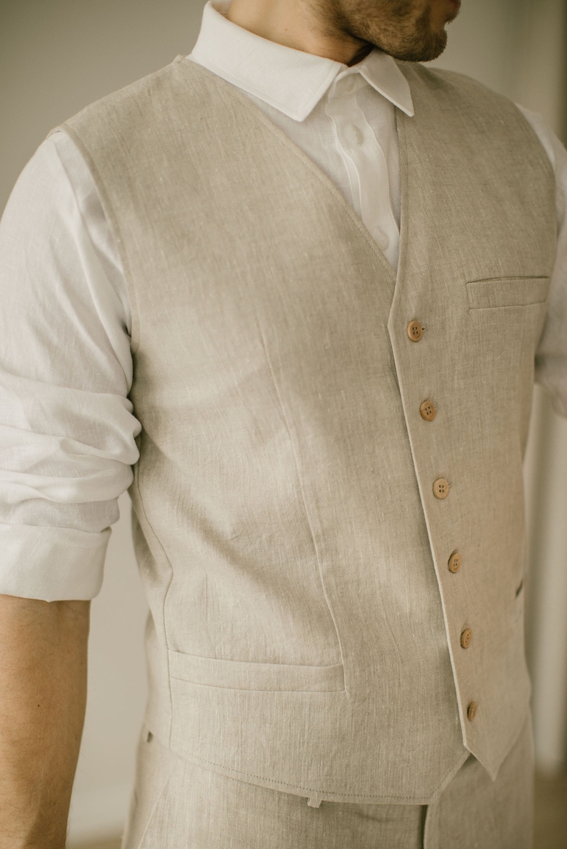 Pu&ograve; includere: Un gilet di lino beige chiaro indossato su una camicia bianca con colletto. Il gilet ha una tasca sul lato sinistro e sei bottoni sul davanti.