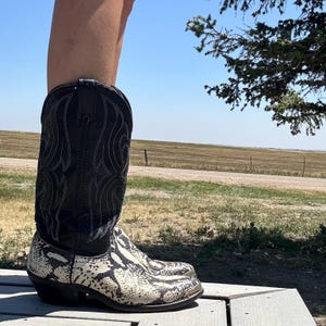 May include: Black and white cowboy boots with a snakeskin pattern on the foot and a black shaft with decorative stitching. The boots are worn outdoors on a wooden deck, with a field and blue sky in the background.
