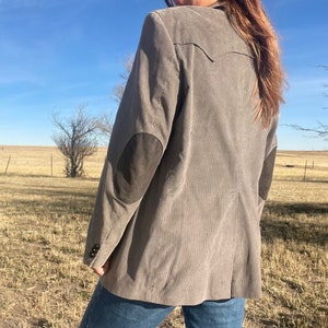 Puede incluir: Una chaqueta de pana gris con parches de cuero marrón en los codos. La chaqueta tiene un cierre de botón único y un ajuste clásico.