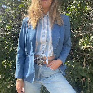 Może przedstawiać: Niebieska kurtka dżinsowa z biało-niebieską paskowaną koszulą pod spodem. Kurtka zapinana jest na jeden guzik i ma srebrną klamrę paska z wzorem dwóch koni. Osoba nosi niebieskie dżinsy.