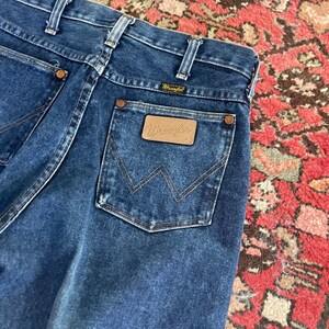 May include: Close-up of dark blue denim Wrangler jeans. The jeans feature a brown leather patch with the Wrangler logo on the back pocket. The stitching is a contrasting tan color. The jeans are folded, showing the back pockets and waistband.