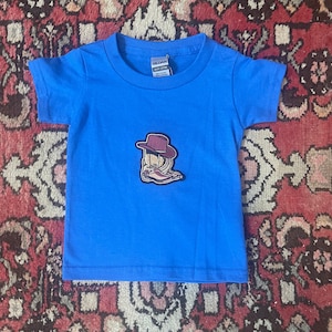 Op de afbeelding: Een felblauw peuter-T-shirt met een cowboy-thema patch. De patch toont een cowboyhoed, laarzen en een touwontwerp. Het shirt is gemaakt van een zacht materiaal en is geschikt voor dagelijks gebruik.