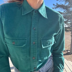 May include: A teal green button-down shirt with two chest pockets and a collar. The shirt has dark buttons and is made of a soft, textured fabric. The shirt is paired with blue jeans.