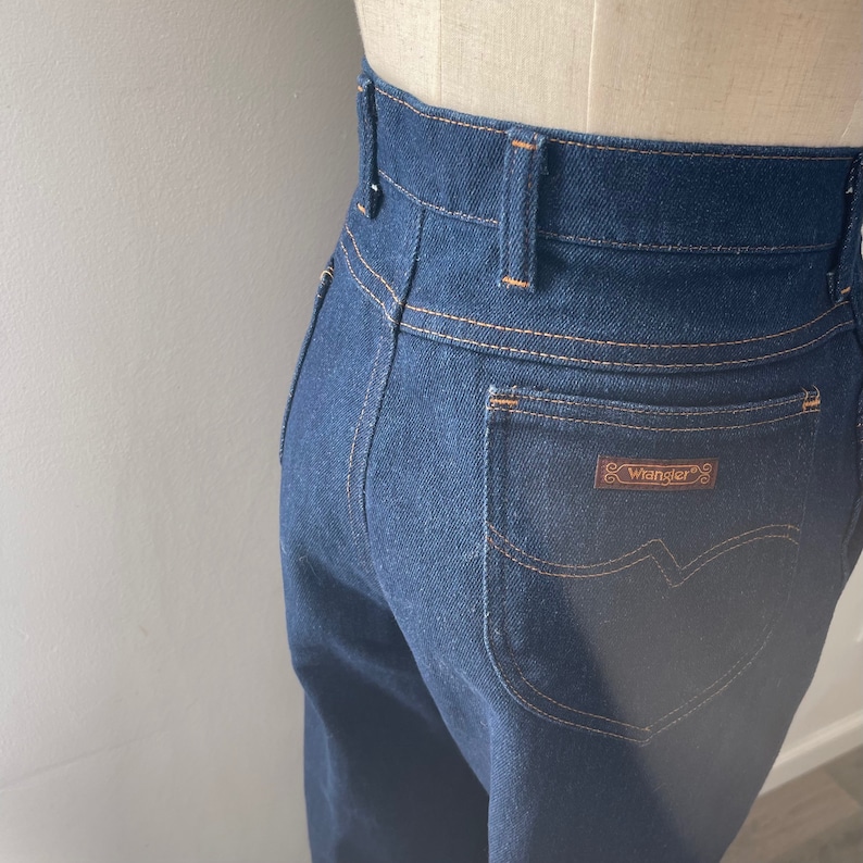 May include: Dark blue denim jeans with a high waist and a back pocket with a brown leather Wrangler label.