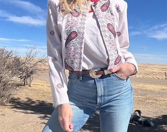 May include: A white cropped jacket with a paisley pattern in shades of red and black. The jacket has a V-neck and long sleeves. The model is wearing blue jeans and a brown belt.