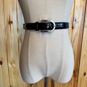 Puede incluir: Un cinturón de cuero negro con hebilla plateada se muestra en un maniquí. El cinturón es de estilo clásico, con un acabado brillante y una hebilla rectangular simple. El maniquí está sobre un soporte metálico.