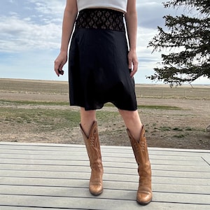 Può includere: Gonna nera con fascia in pizzo e stivali da cowboy marroni. La gonna è lunga fino al ginocchio e sembra essere realizzata in un tessuto leggero. Gli stivali sono di colore marrone chiaro e hanno un classico design western.