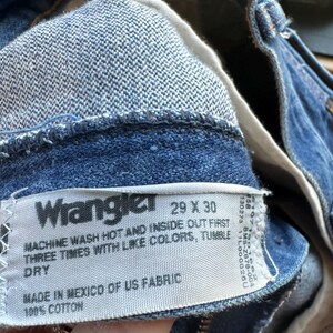 May include: Close-up of blue denim Wrangler jeans. The label reads "Wrangler 29 X 30" and includes washing instructions. Made in Mexico of US fabric, 100% cotton. The jeans are a classic style.
