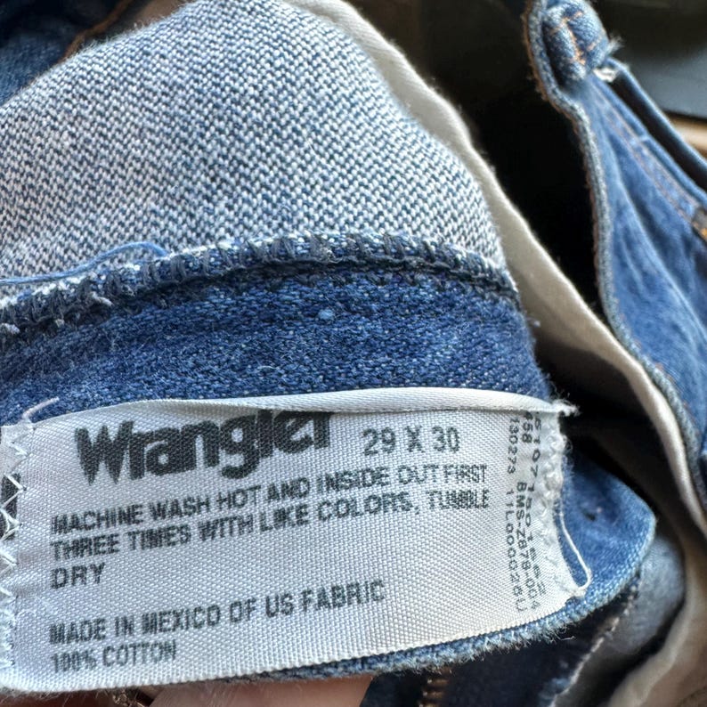 May include: Close-up of blue denim Wrangler jeans. The label reads "Wrangler 29 X 30" and includes washing instructions. Made in Mexico of US fabric, 100% cotton. The jeans are a classic style.