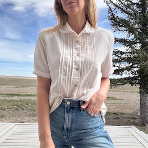 May include: A short-sleeved cream-coloured button-down blouse featuring vertical pintuck pleats along the front placket, a rounded collar with delicate white embroidery, and matching lace trim detailing on the sleeve cuffs and collar edges.