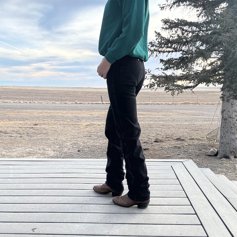 May include: A person wearing a dark green sweatshirt and black jeans stands on a wooden deck. The person is also wearing brown cowboy boots. The background includes a field, a road, and a cloudy sky.
