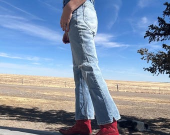 Puede incluir: Una persona lleva unos vaqueros de mezclilla azul claro con un detalle desgastado en la pierna izquierda y botas de vaquero rojas. La persona está de pie sobre una superficie de madera con un cielo azul y un campo marrón en el fondo.