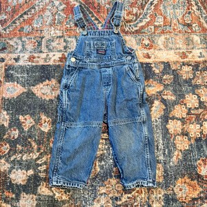May include: Blue denim overalls with adjustable straps and multiple pockets. The bib features a small patch with text. The overalls have a classic design, suitable for children's wear.
