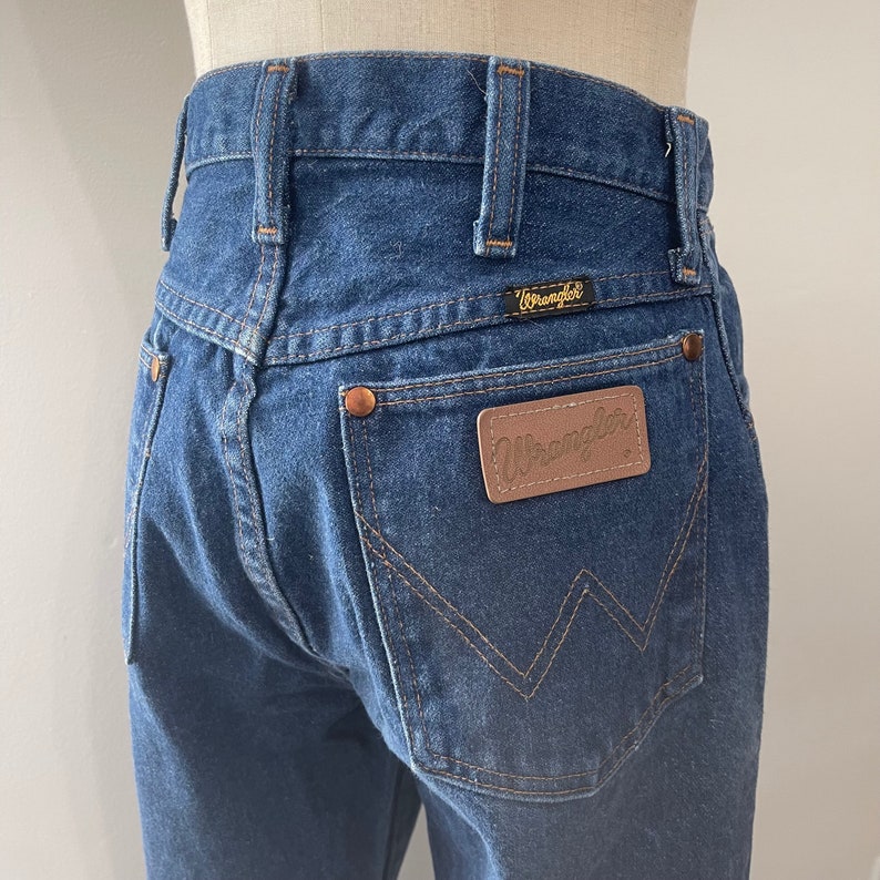 Pu&ograve; includere: Un paio di jeans blu denim con una toppa in pelle marrone sulla tasca posteriore che dice "Wrangler".