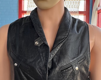 May include: A black leather vest featuring a wide collar, a front snap button closure, and two horizontal zippered chest pockets with silver-toned hardware. The vest has a subtle quilted pattern across the surface and a structured, sleeveless design.