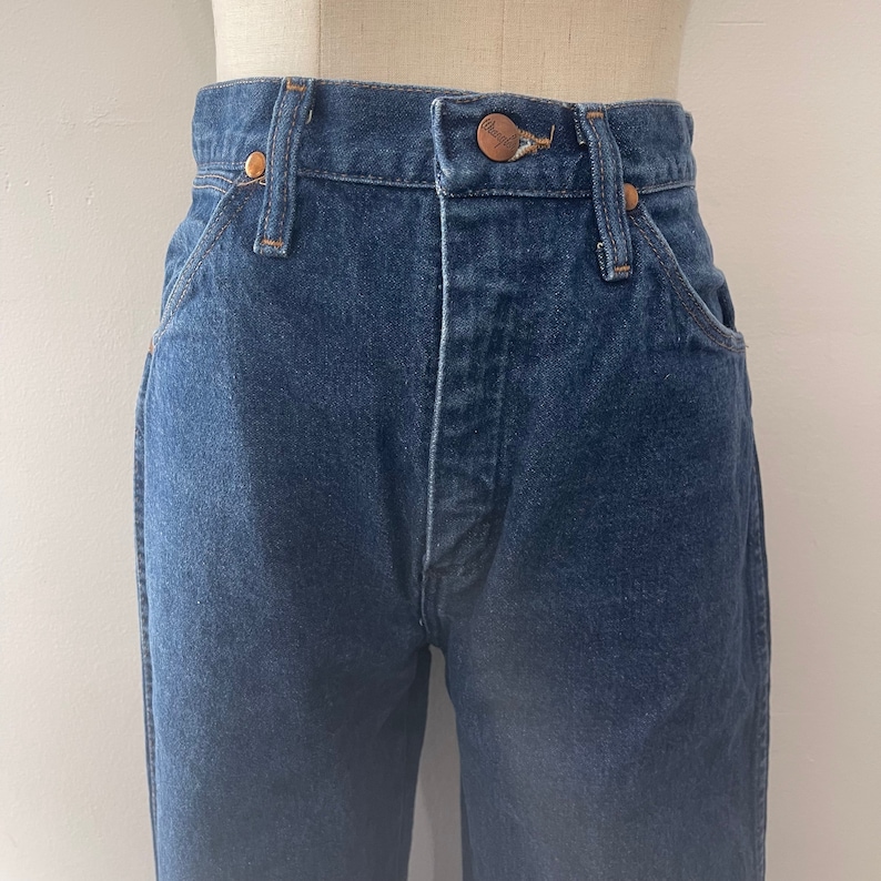 Pu&ograve; includere: Un paio di jeans blu denim con un bottone e rivetti in rame. I jeans hanno vita alta e gamba dritta.