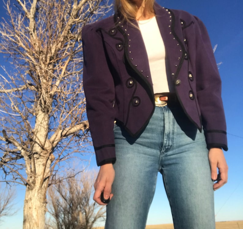 May include: A purple cropped jacket with black trim and silver buttons. The jacket has a pointed collar and a fitted silhouette.