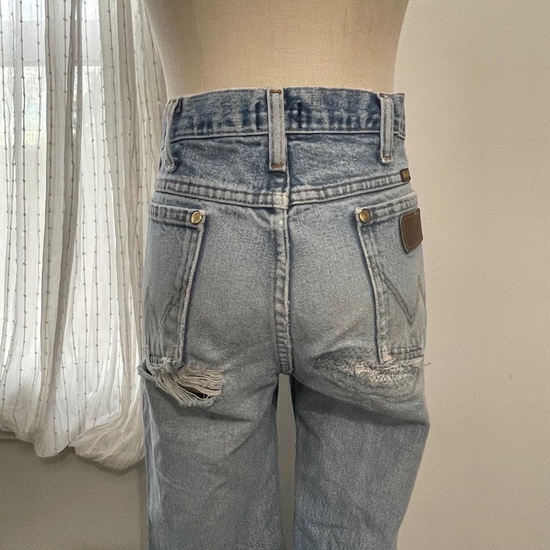 May include: Light blue denim jeans with a distressed look. The jeans have a high waist and a straight leg. The back pockets have a brown leather patch with the Wrangler logo.