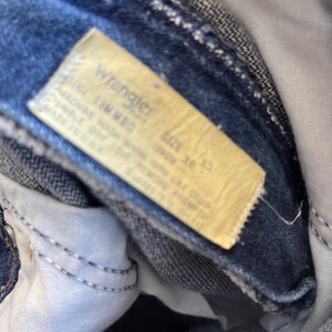 May include: Close-up of a pair of blue denim jeans with a yellowed tag. The tag reads "Wrangler" and includes size information: 36 x 33. The jeans are folded, showing the inside pocket and stitching.